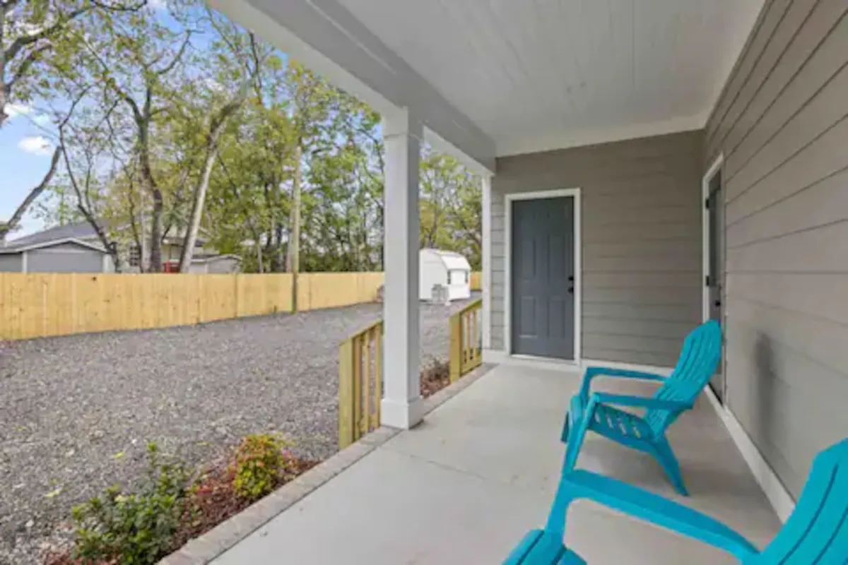 A bright, casual exterior with blue chairs positioned on the porch, a front door, and a gravel driveway framed by leafy trees. The outdoor setting suggests a tranquil retreat that feels distinct yet connected to the urban setting, with space for gathering or enjoying quiet moments.