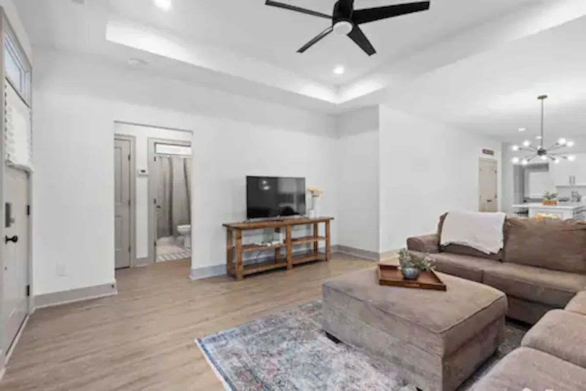 A bright, inviting living room with a comfortable sofa arranged around a TV stand, a patterned area rug, and a ceiling fan above. Natural light streams in, suggesting a relaxing, shared space perfect for families to gather after exploring the city or attending campus events.