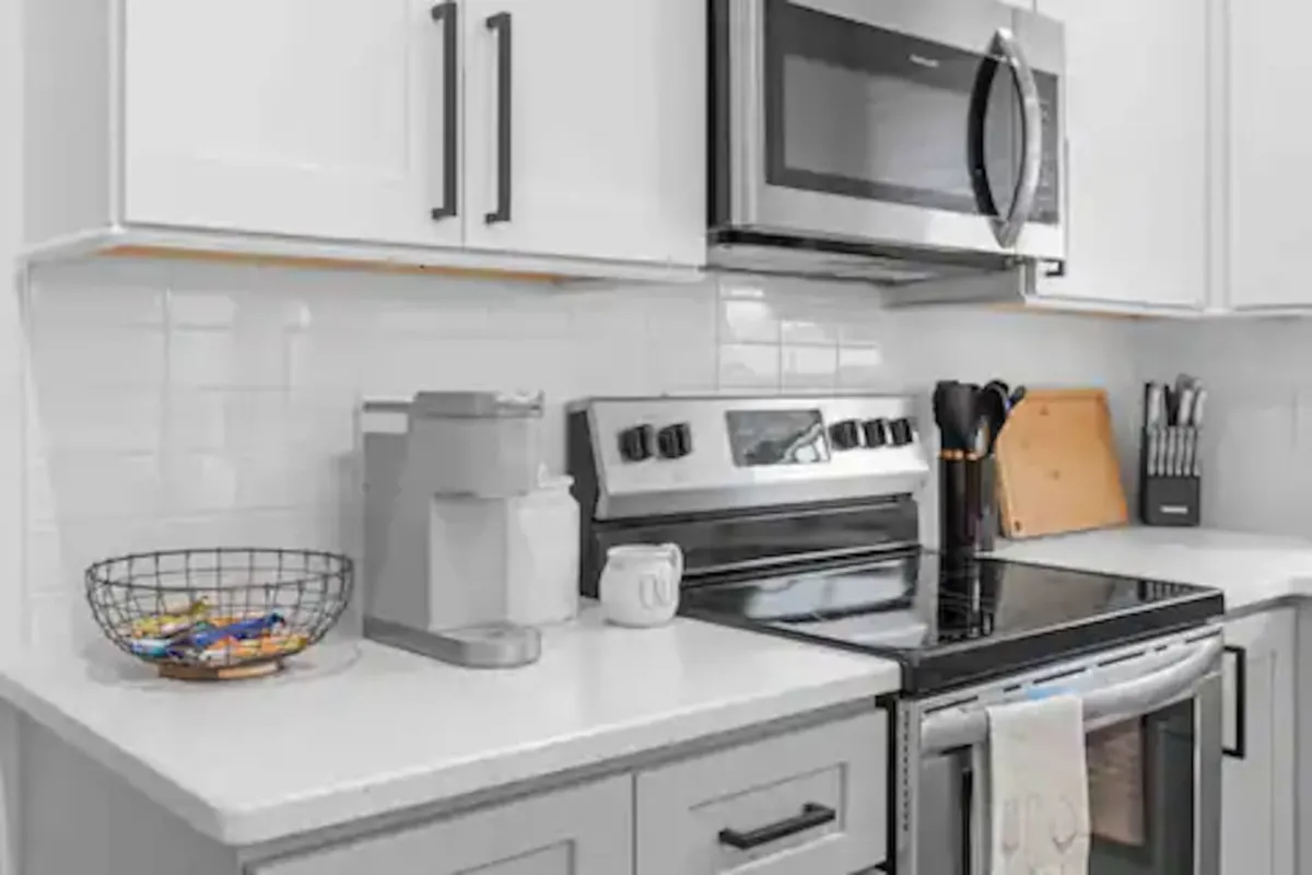 A functional kitchen showcasing stainless steel appliances, white cabinetry, black accents, and thoughtful details like kitchen utensils and a basket of snacks. The space is organized and ready for cooking, baking, or casual get-togethers.