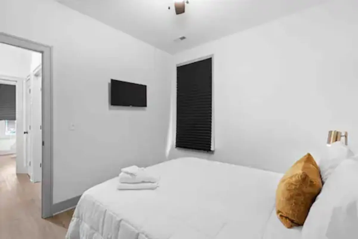 A serene bedroom featuring a large bed with crisp white linens and a golden pillow, complemented by a wall-mounted TV. The room exudes a calm atmosphere ideal for rest and relaxation, with modern touches and uncluttered design.