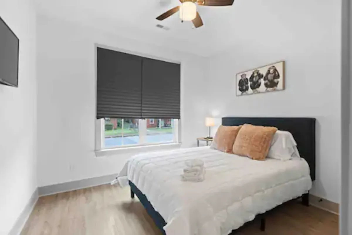 A warm, inviting bedroom with a neatly made bed positioned near a large window, soft lighting from a table lamp, and tasteful wall art creating a peaceful ambiance. The setting feels cozy and restful, ideal for relaxation after a day in the city.