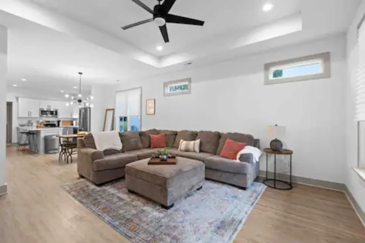 This image shows a spacious open-concept living area with a large sectional sofa and coffee table, accented by a decorative rug and eye-catching artwork on the walls. The kitchen area is visible in the background, showcasing the fluid connection between living, dining, and cooking spaces. Natural light pours in, making the room bright and inviting, perfect for gatherings and relaxation.