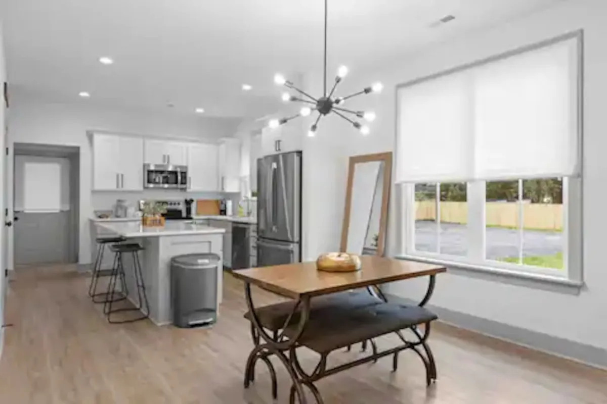 A cozy living-dining space featuring a modern light fixture, a wooden dining table ready for shared meals, a comfortable sofa, and a sleek television unit. Large windows allow sunlight to stream in, highlighting both the dining and lounging areas, creating a lively yet relaxing environment.