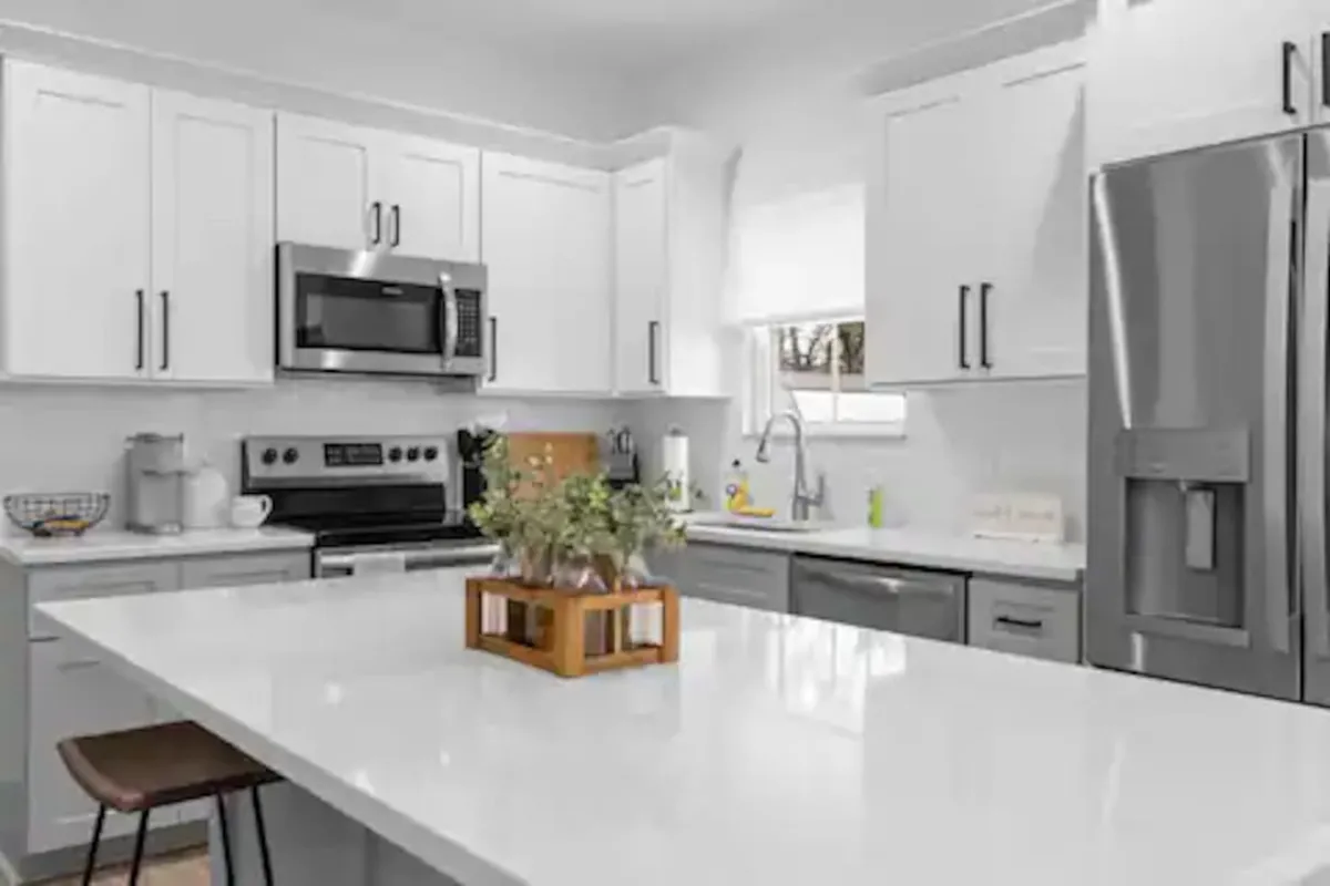 A bright, inviting kitchen featuring a central island, gleaming granite countertops, modern stainless steel appliances, white cabinetry, and a decorative vase with fresh flowers. Ample natural light streams in, highlighting the spacious and functional layout that makes both meal prep and socializing easy.