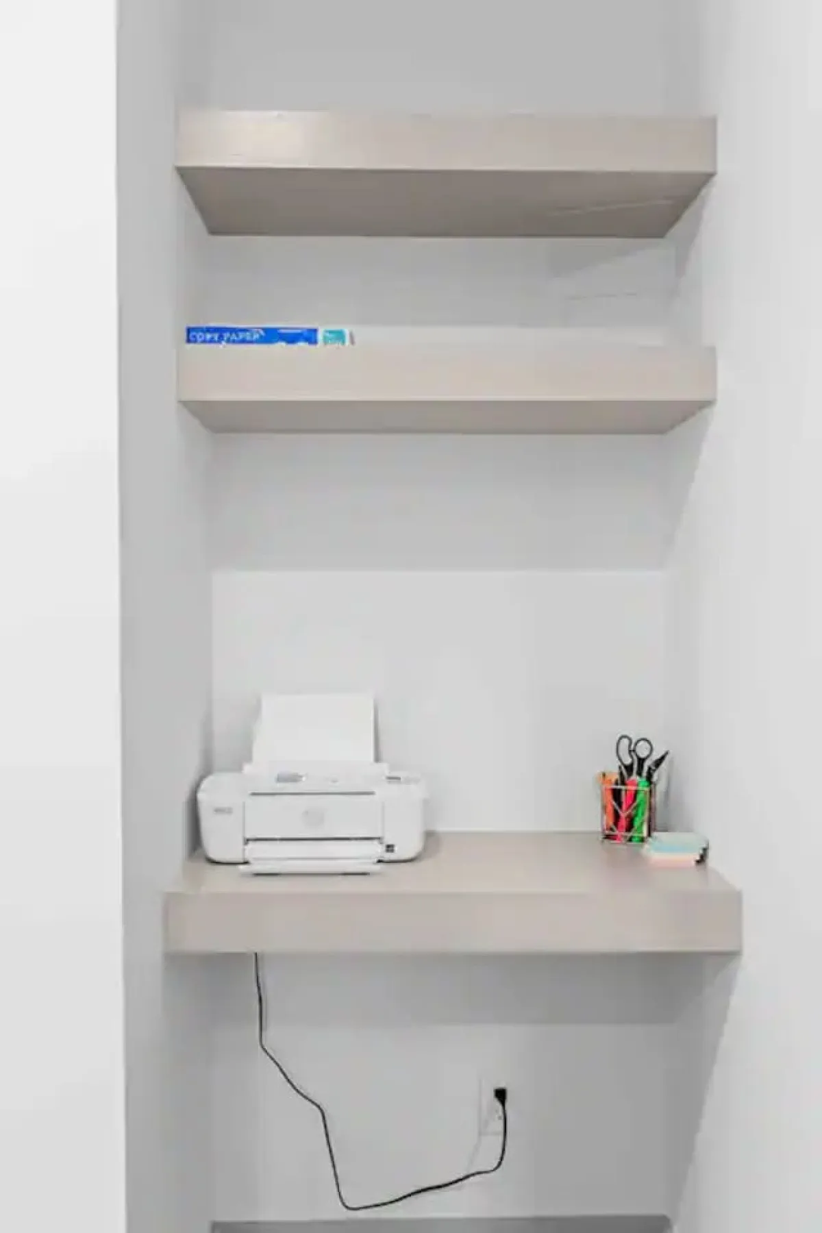 This image depicts a minimalistic and organized workspace with a printer, shelves, and stationery supplies. The setup suggests an efficient area for handling work-related documents or staying productive while working remotely.